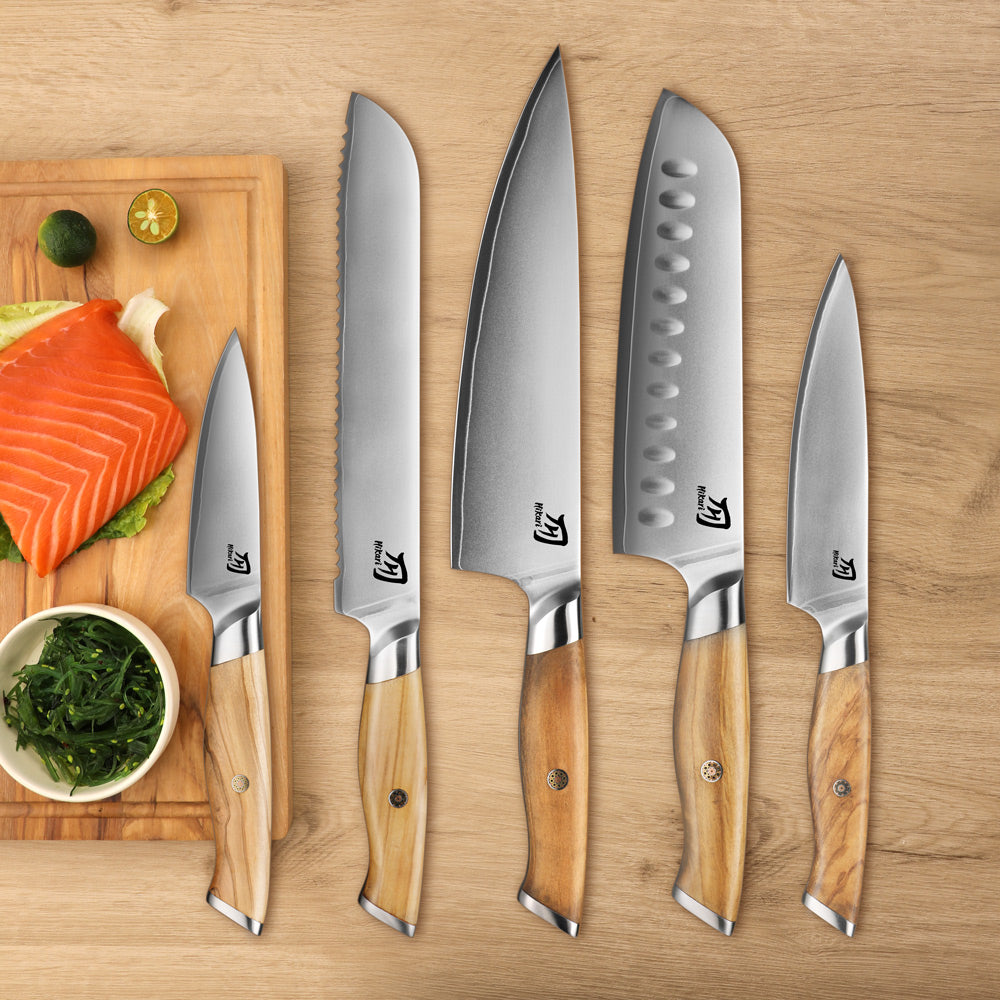 Five kitchen knives with light wood handles are arranged on a wooden surface beside a cutting board with salmon, lime, and greens. The knives have silver stainless steel blades and are branded with a stylized 'K' logo.