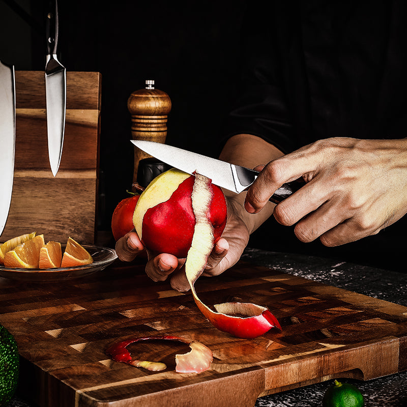 Person peeling a red apple on a wooden cutting board with a knife and other fruits in the background.
