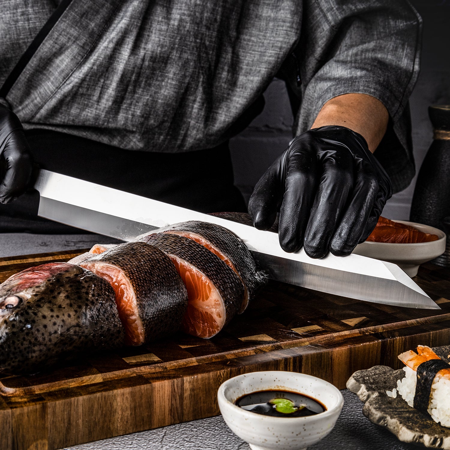 Person in black gloves cutting raw salmon with a premium fish knife branded Hikari Knife called Yanagiba japanese knife on a wooden board, surrounded by sushi ingredients.